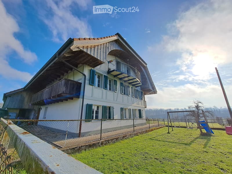 Two-story house with white walls, green shutters, and wooden balconies. The house is surrounded by a fenced yard with a swing set and grassy area.