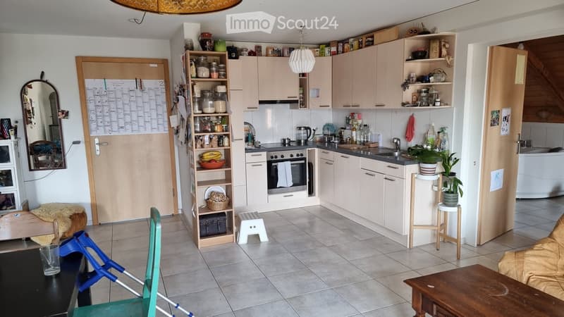 A cozy kitchen and living area with light-colored cabinets, a central island, a sofa, a table, and various kitchen items on shelves and counters.