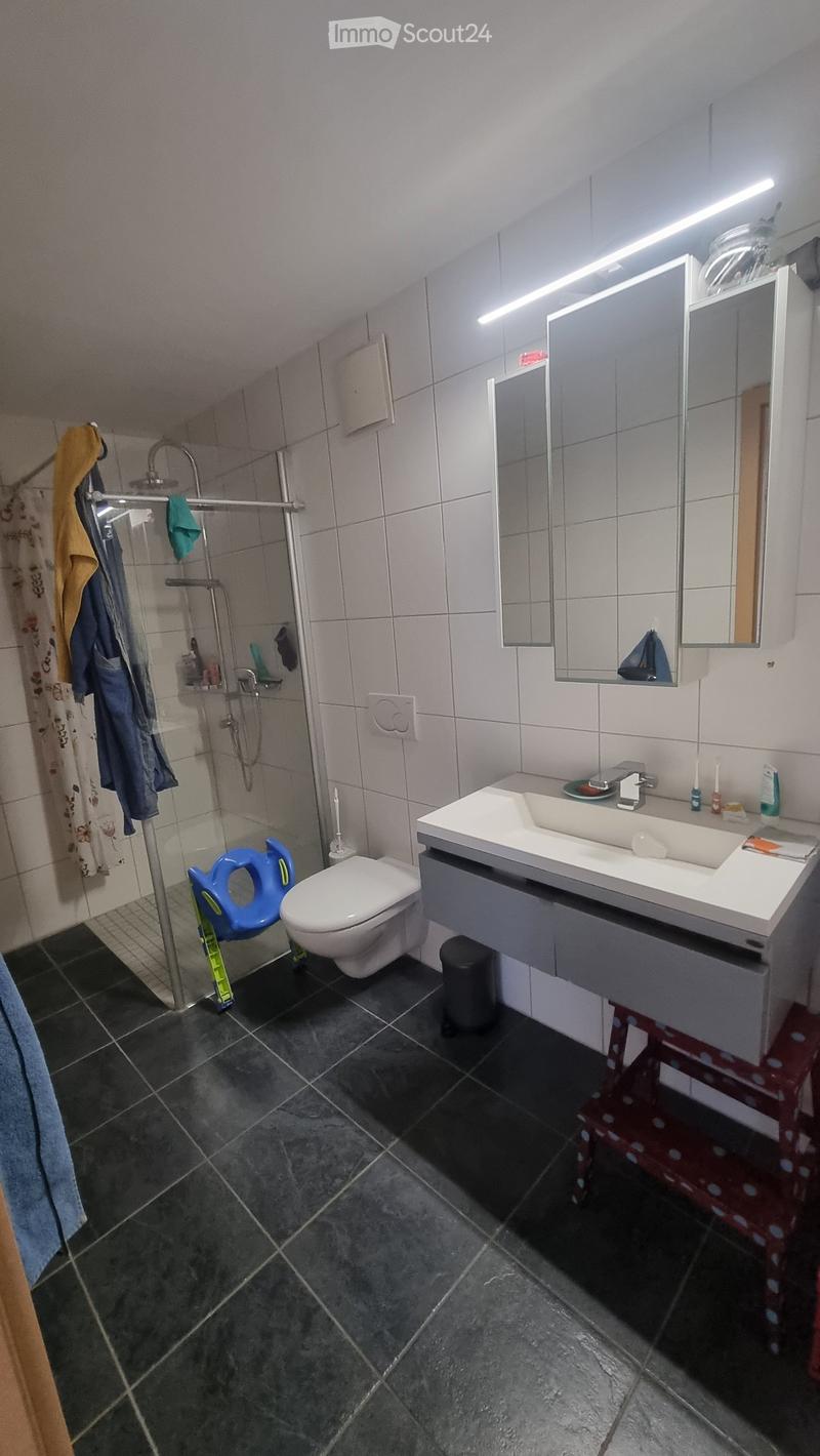 Modern bathroom with white tiles, dark grey floor, large mirror, sink, toilet, and shower area with towel rack and stool.