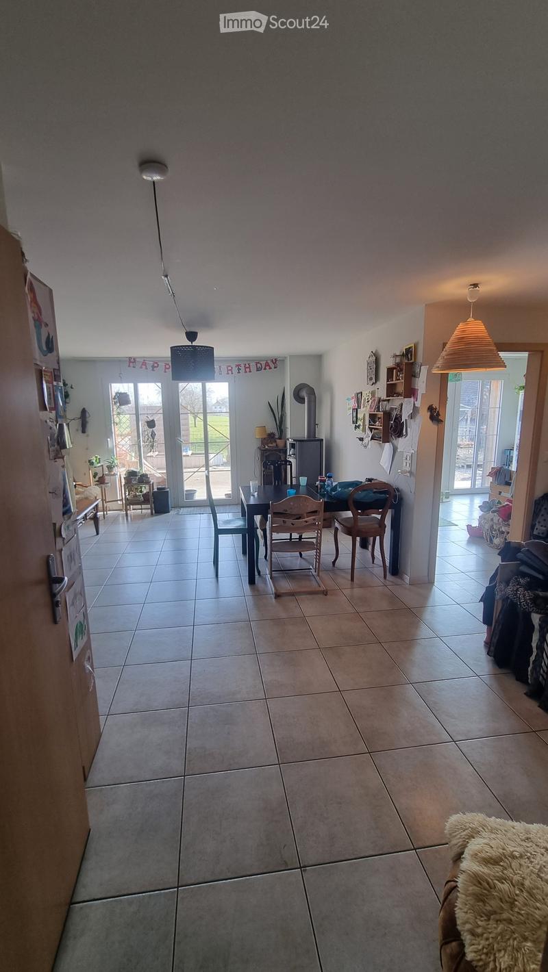 Bright living area with tiled floor, large windows, and a dining table with chairs. A refrigerator is visible on the left, and a bench with a fur throw is in the foreground.