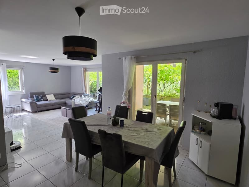 Bright living and dining area with a grey sectional sofa, coffee table, dining table with chairs, and a small cabinet with a coffee maker. Large windows and sliding glass doors lead to a balcony.