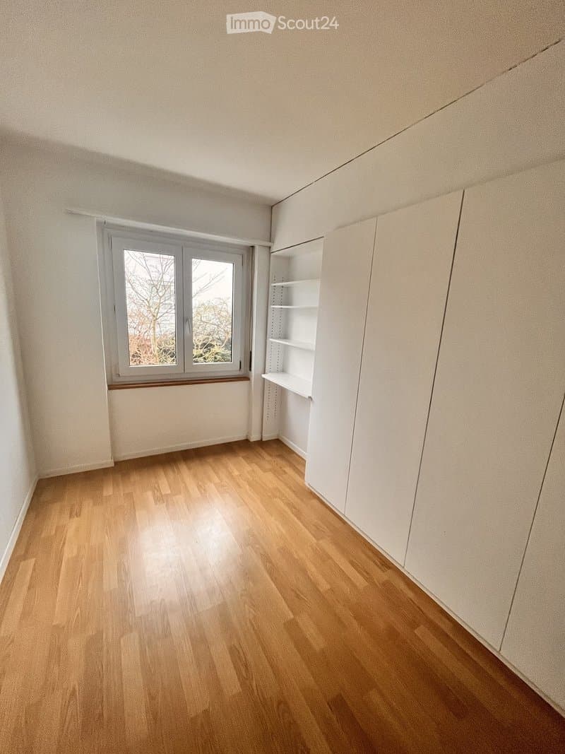 Empty room with wooden flooring, white walls, built-in shelving unit, and a window with white frames.