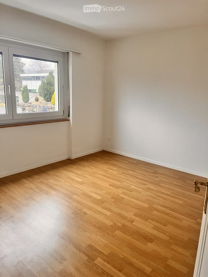 Empty room with light wooden flooring and white walls, featuring a large window with a view of greenery outside.