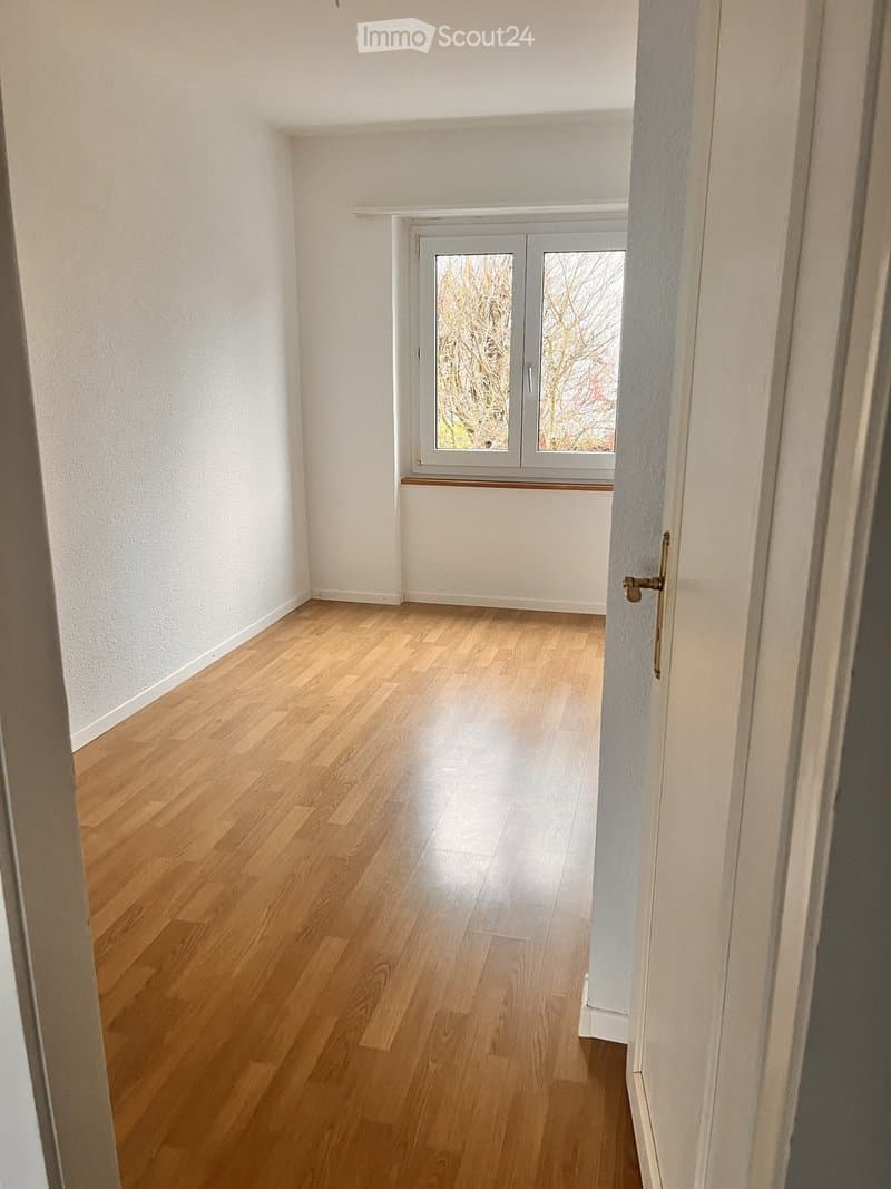 Empty room with light wooden flooring and white walls, featuring a window with double glass panels and a simple door frame.