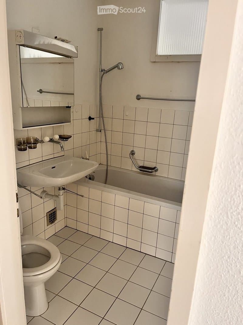 A small bathroom with white tiled walls and floor, featuring a toilet, sink, bathtub, mirror, and a handrail for safety.
