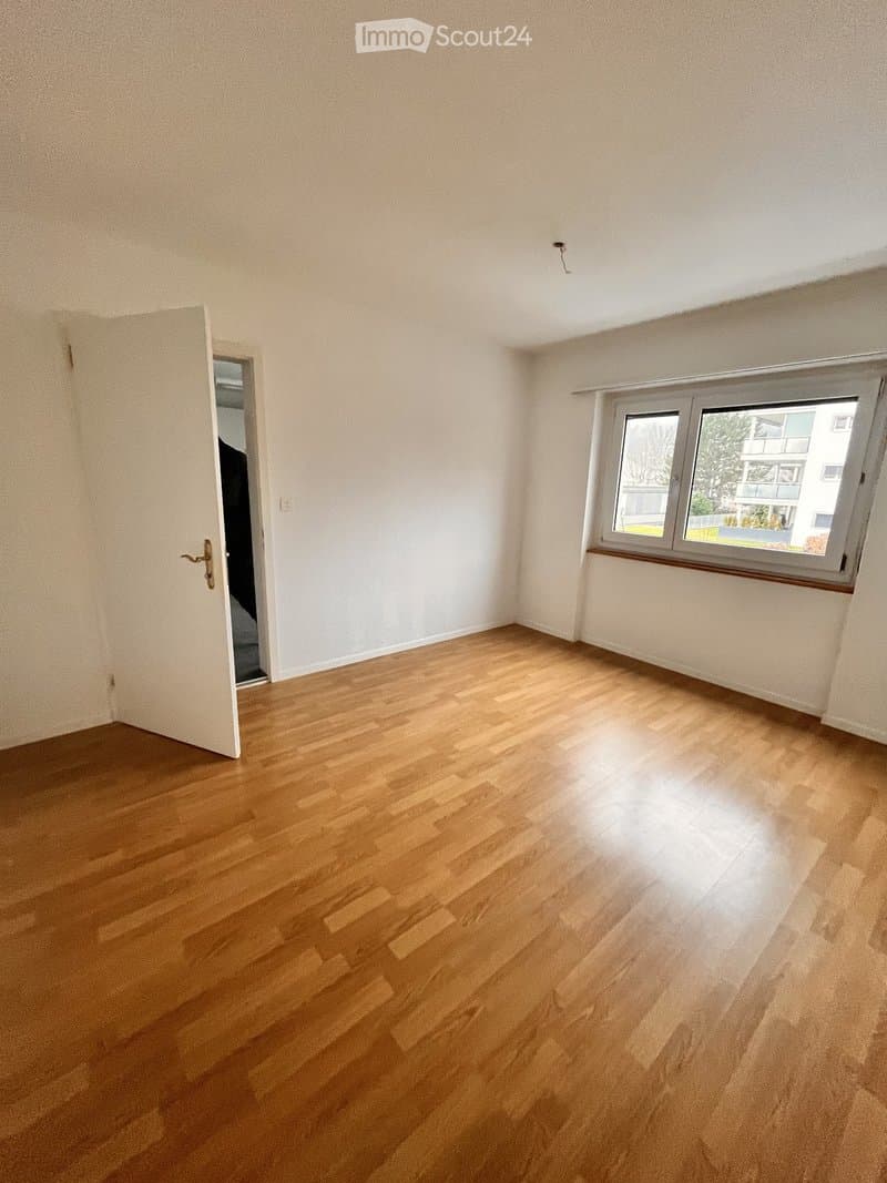 Empty room with light wooden flooring, white walls, and a large window with a view of the outside. The room has a door leading to another space.