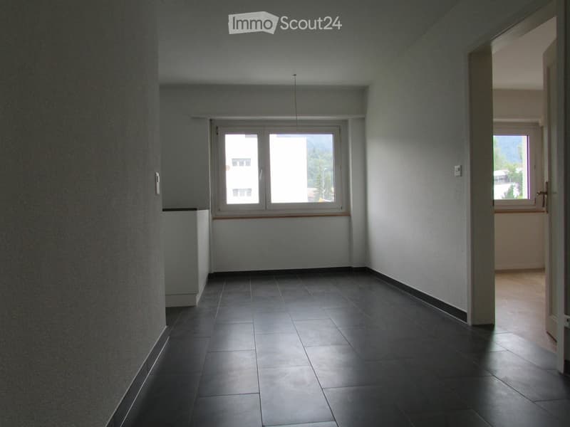 Empty hallway with tiled floor, white walls, and large windows at one end, leading to another room.