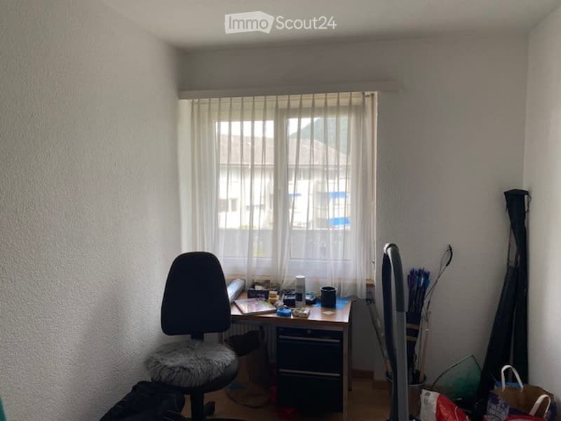 A small, furnished room with a desk, chair, and various items on the desk including a laptop, cup, and lamp. There is a window with white curtains and some bags and cleaning supplies near the wall.