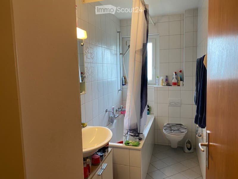 Bathroom with white tiled walls and floor, bathtub with shower curtain, toilet, sink, and various cleaning supplies on the shelf.