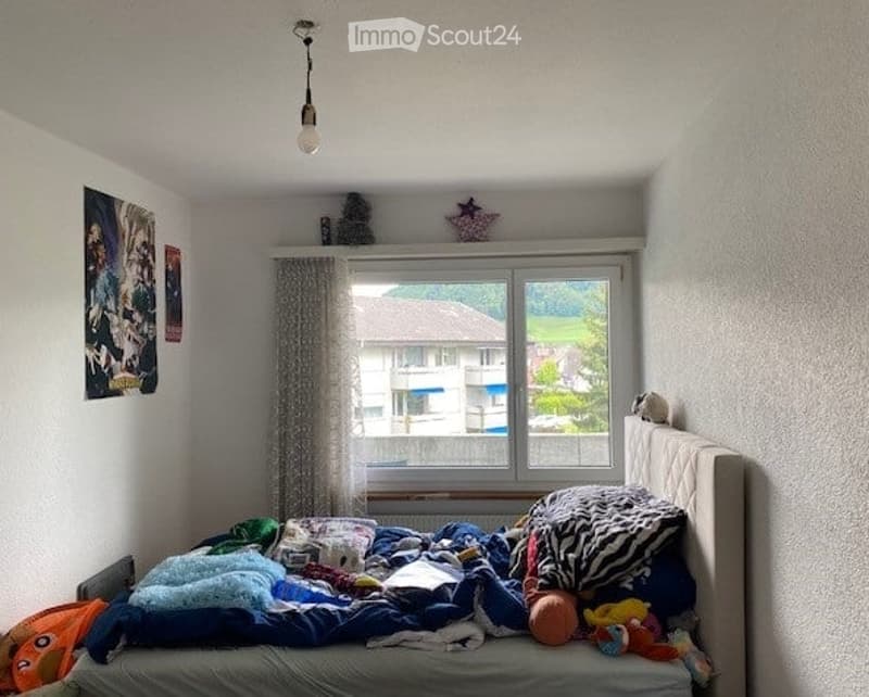 A small bedroom with a bed covered in blankets and pillows, a window with a curtain, and a picture frame on the wall. The room has a simple, cozy feel with a view of a neighboring building outside.