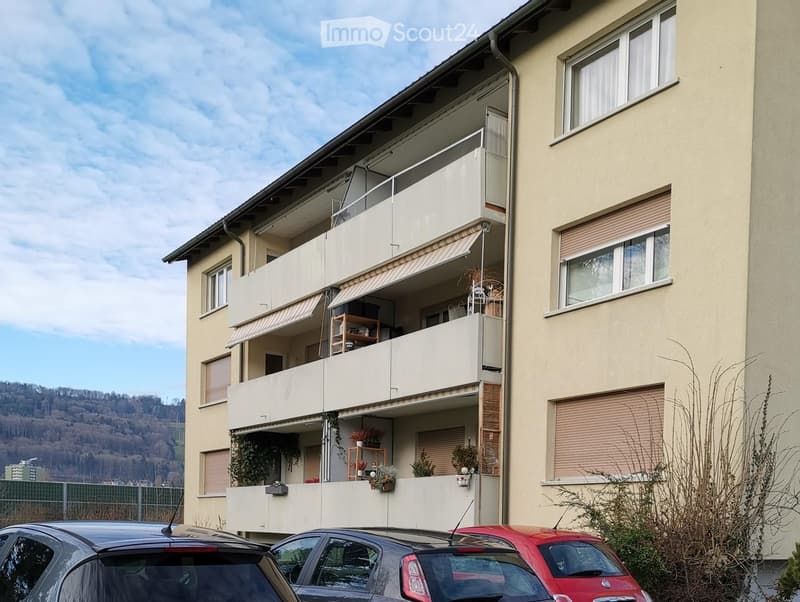 Multi-story residential building with balconies, windows, and awnings. Several cars are parked in front.