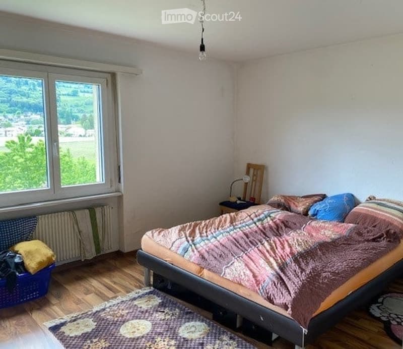 Bedroom with wooden flooring, a large window with a scenic view, a bed with colorful bedding, a nightstand with a lamp, a small rug, and a basket on the floor.