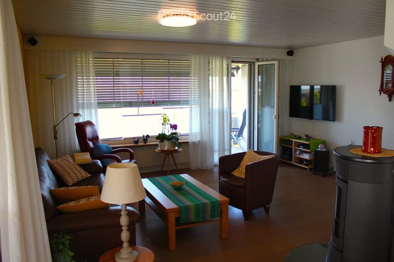 Bright living room with wooden flooring, large window with blinds, sofa, armchair, coffee table, table lamp, floor lamp, television, side table with potted plant, and sideboard.
