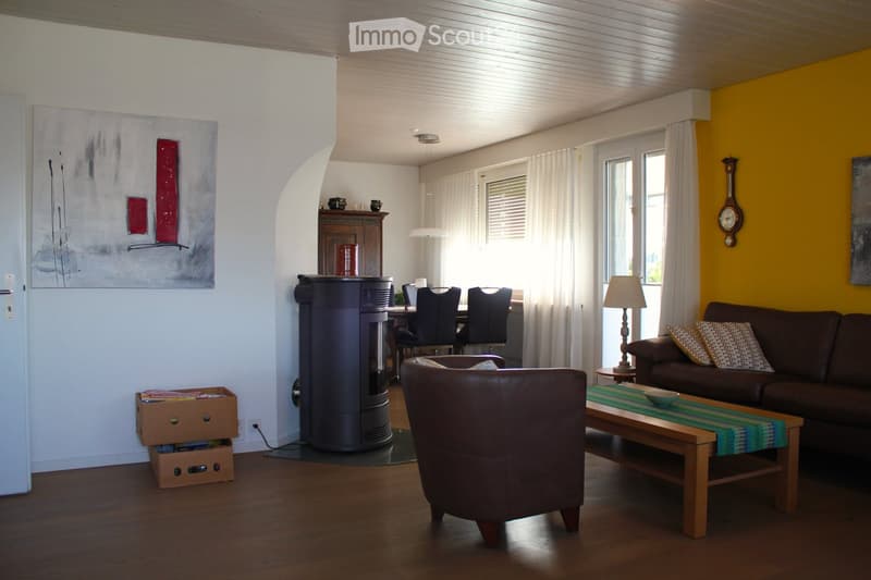 Living room with wooden flooring, a brown leather armchair, a sofa with patterned cushions, a coffee table with a green runner, a fireplace, a dining area with a table and chairs, and a lamp on a side table.