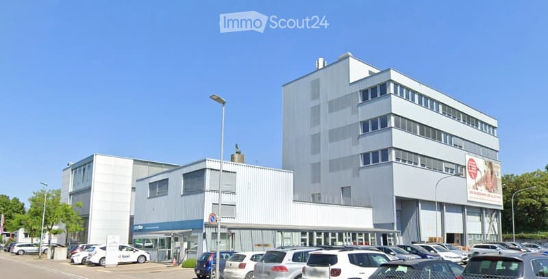 Modern multi-story building with a combination of glass and metal facade, surrounded by a parking lot with numerous cars.