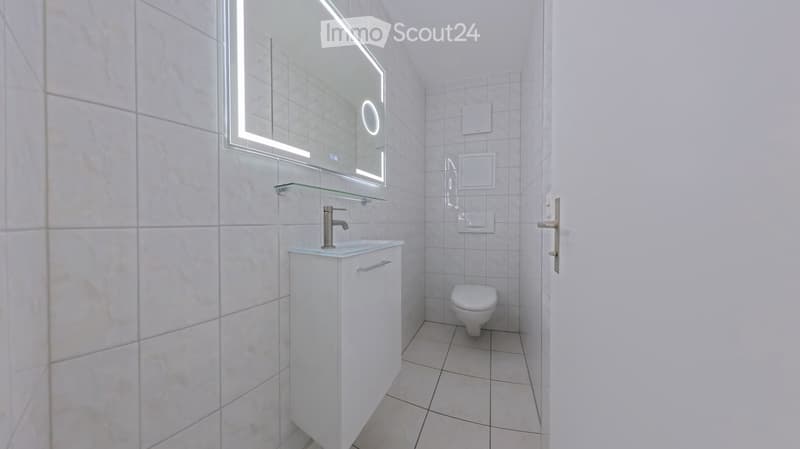 Modern bathroom with white tiles, a sink with a sleek faucet, a large illuminated mirror, and a toilet.