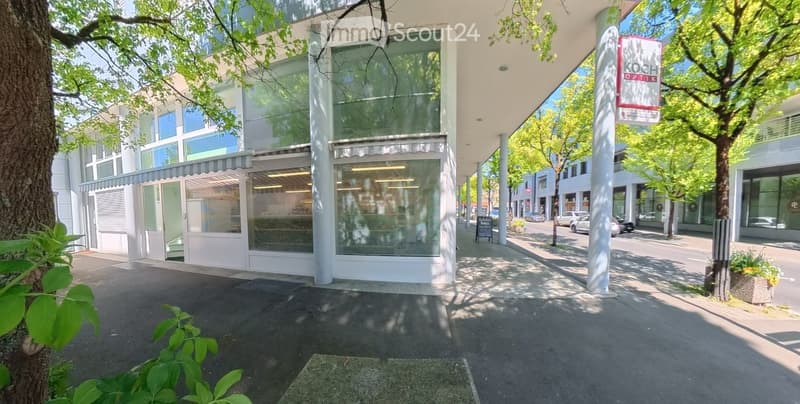 Modern building with glass windows, awnings, and a signboard. Trees and plants are in front of the building. A car is parked on the side of the road.