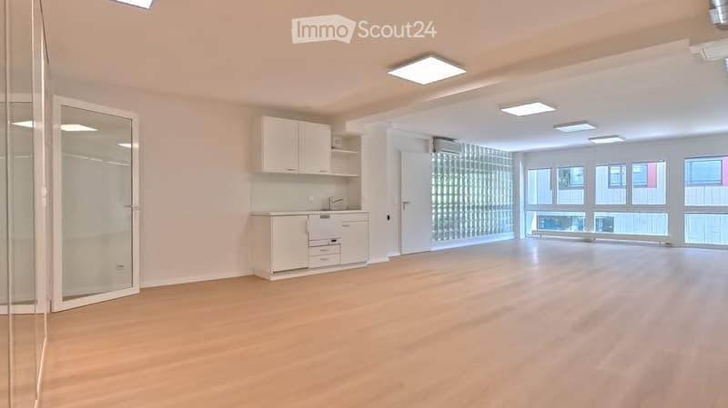 Empty room with light wooden flooring, white walls, and a small kitchenette area with cabinets and a sink. Large windows provide natural light.