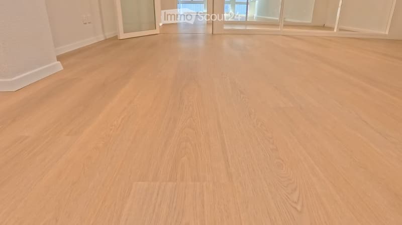 Empty room with light wooden flooring, white walls, and large glass doors leading to another space.