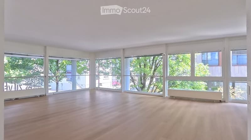 Empty room with wooden flooring and large windows offering a view of greenery outside.