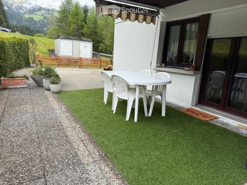 Outdoor patio with artificial grass, white plastic dining set, white house with large windows, scenic mountain view in the background.