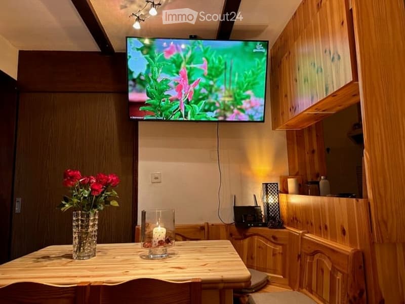 A cozy, wooden-paneled room with a flat-screen TV mounted on the wall, a small wooden table with a vase of red flowers and a candle, and wooden chairs around the table. Wooden cabinets line the right side of the room.