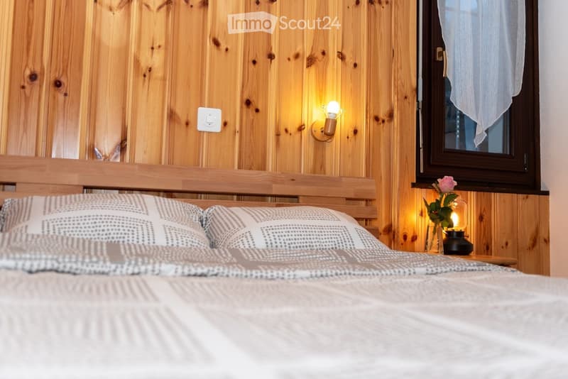 Cozy bedroom with wooden walls, a neatly made bed with patterned pillows and a textured duvet, a nightstand with a vase of flowers and a small lamp, and a wall-mounted light fixture providing warm illumination.