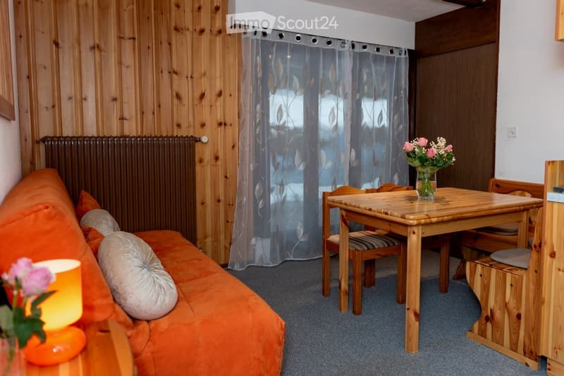 Cozy living room with a wooden table, sofa, and chairs. Features a radiator, wooden walls, and a curtained window.