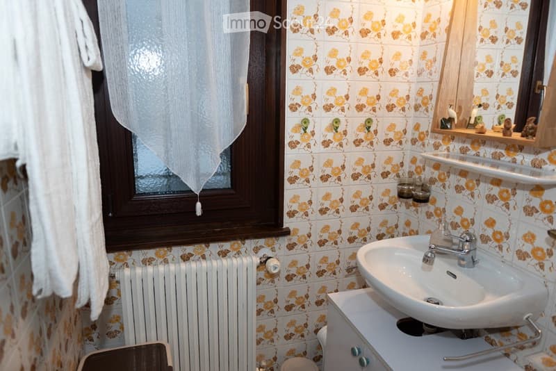 Bathroom with floral-patterned tiles, a white sink with a chrome faucet, a radiator, a mirror with shelves, and a towel hanging on the wall.