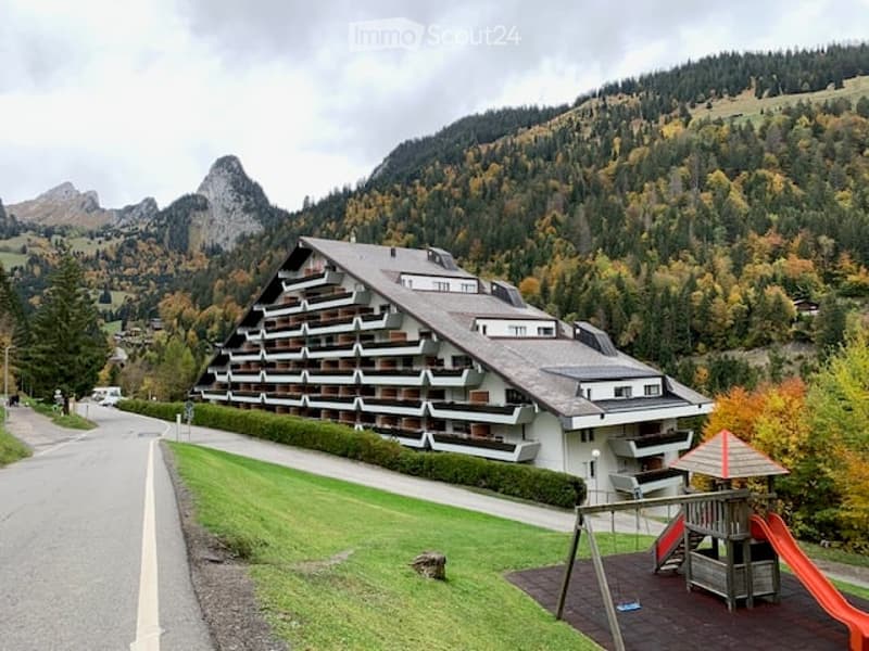 A multi-story apartment building with balconies, situated in a scenic mountainous area with a playground in the foreground.
