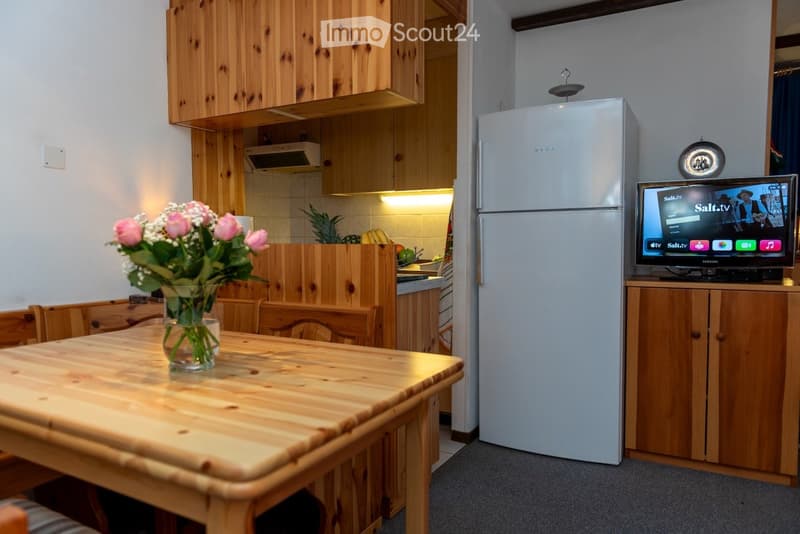 A cozy, well-furnished room with a wooden dining table and chairs, a vase with pink flowers, a refrigerator, and a television on a wooden stand. The kitchen area features wooden cabinets and a countertop with fruit and vegetables.