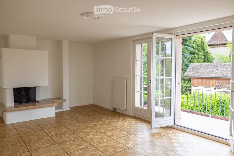 Empty room with wooden flooring, fireplace, large sliding glass doors leading to a balcony, and a view of greenery and a neighboring building.
