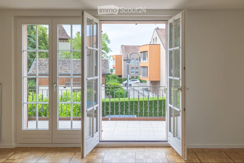 Open sliding doors lead to a tiled balcony with a metal railing, overlooking a residential area with houses and greenery.