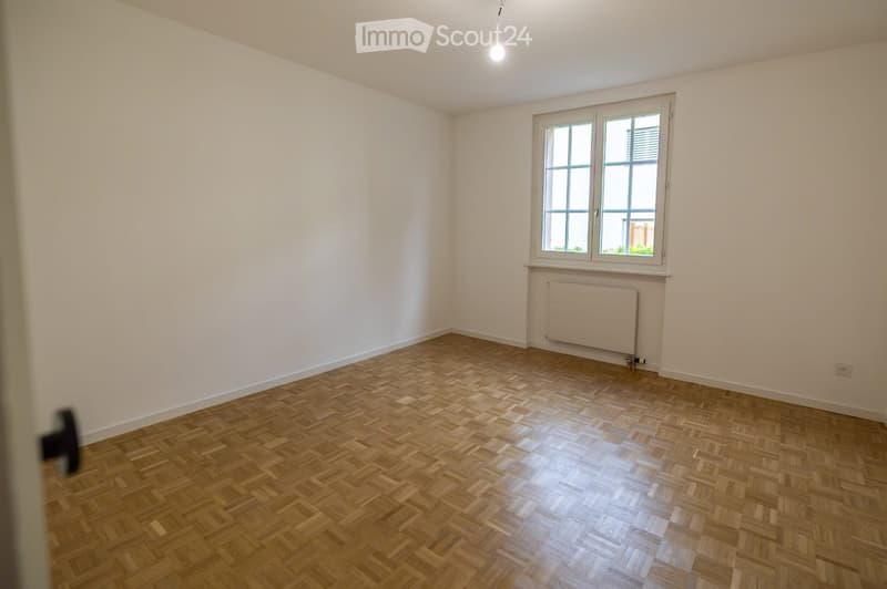 Empty room with wooden parquet flooring, white walls, a window with white frame, and a radiator below the window.
