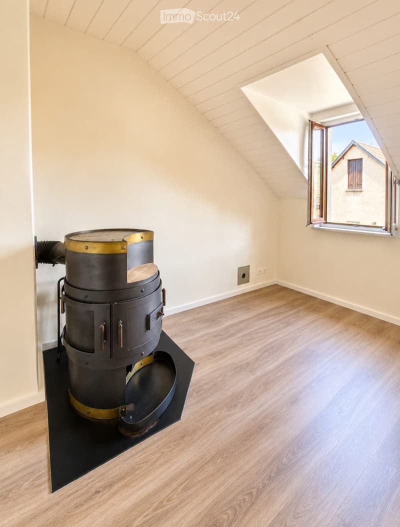 An empty attic room with wooden flooring, white walls, and a skylight window. A large, industrial-style stove is positioned against the wall.
