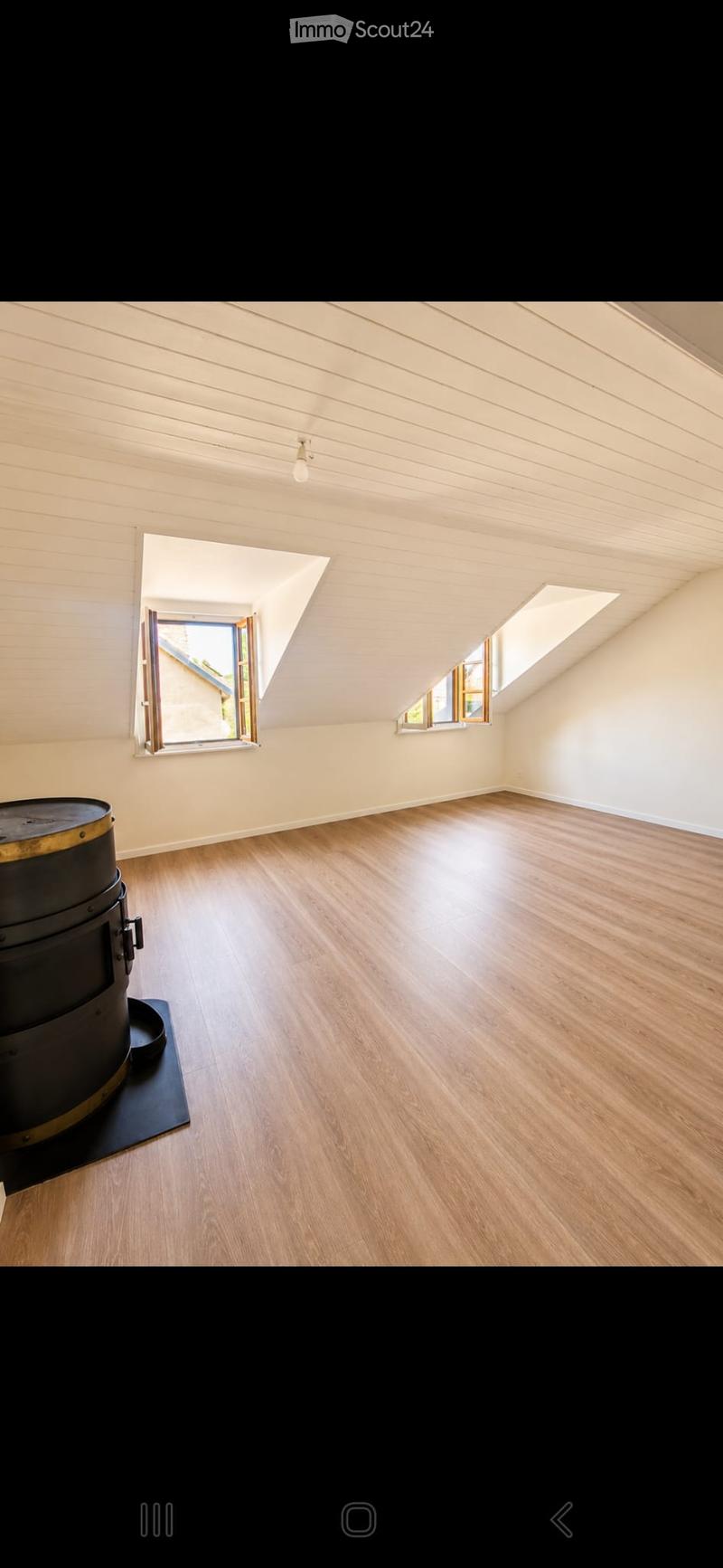 Empty attic room with wooden flooring, slanted wooden ceiling, and multiple windows allowing natural light. A black cylindrical object is placed on the floor near the left side.