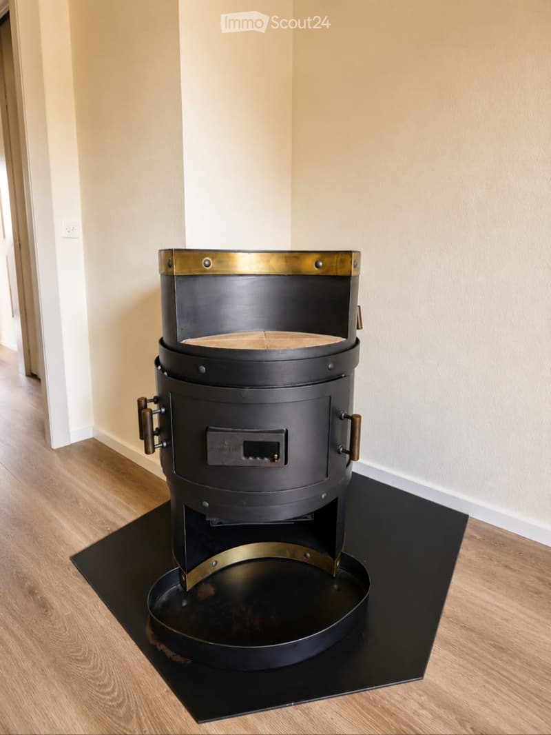 A modern hallway with light-colored walls and wooden flooring, featuring a black and gold cylindrical fireplace in the center.