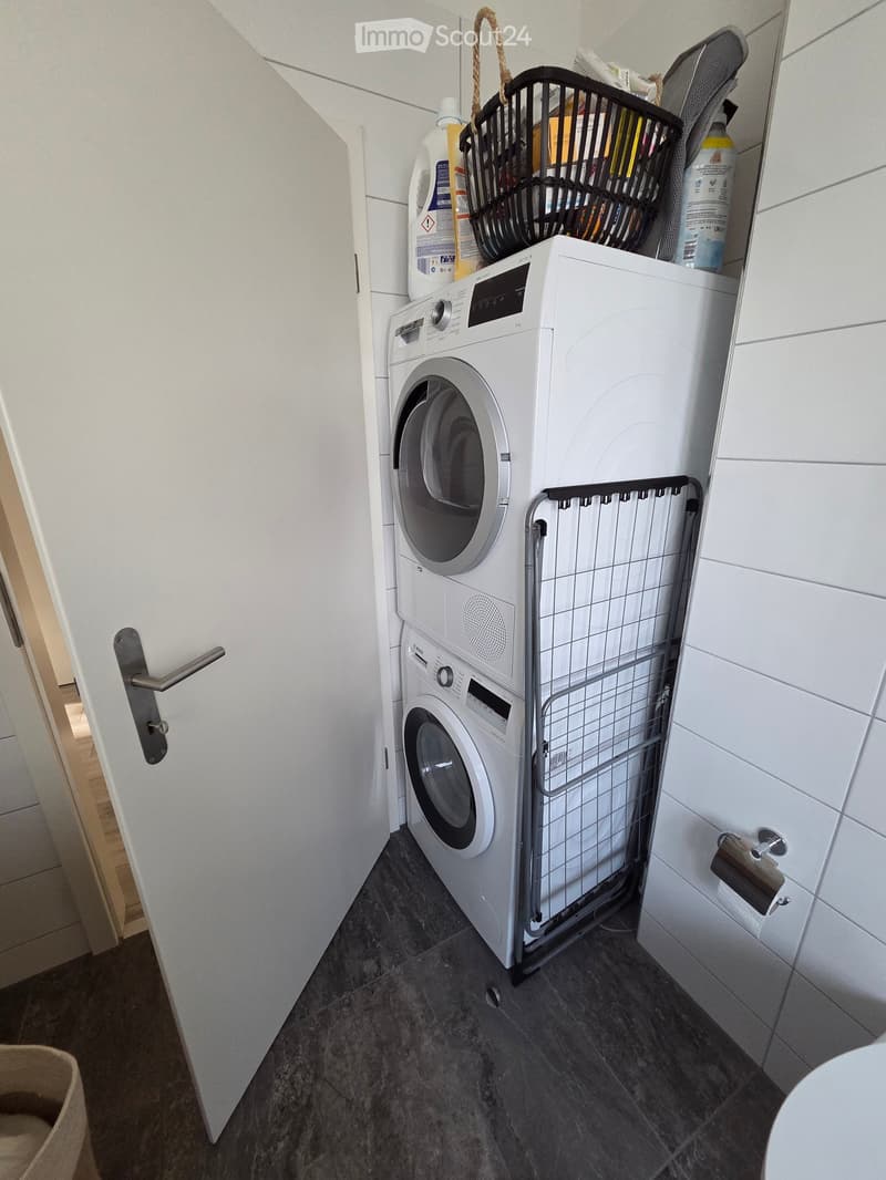 Laundry area with a washing machine, dryer, and a laundry basket on top. Dark tiled floor and white tiled walls.