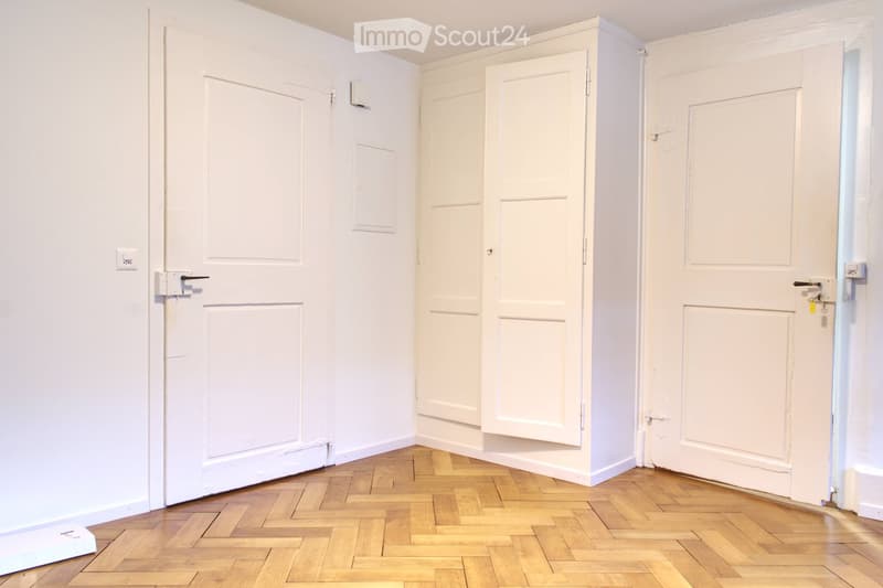 Empty room with white walls, wooden parquet floor, three doors, and built-in cabinets.