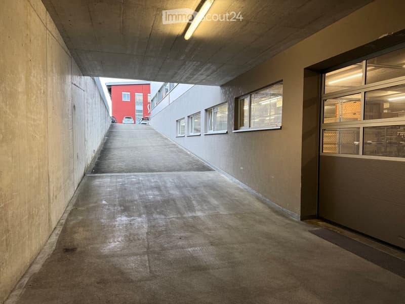 A concrete-covered corridor with a smooth floor, walls, and windows on one side, leading to an open area with visible buildings and parked cars.