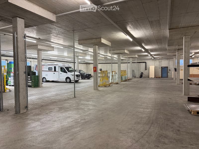 Underground parking garage with concrete floors and pillars, featuring a fenced area with a van and other vehicles, storage containers, and a blue door.