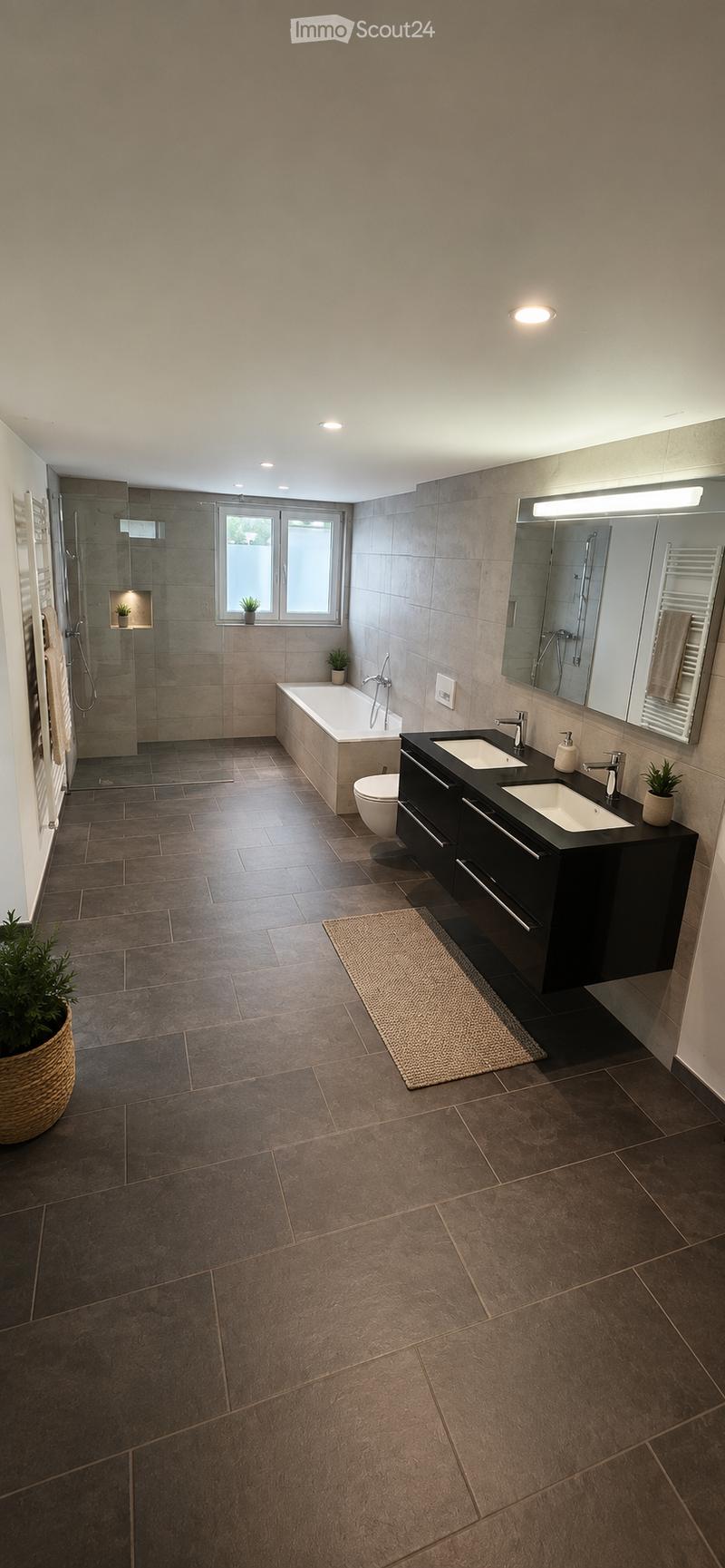 Modern bathroom with dark tiled floors, a large bathtub, a double sink vanity, a shower area, and a large mirror with built-in lighting. Includes a small potted plant and a woven rug.