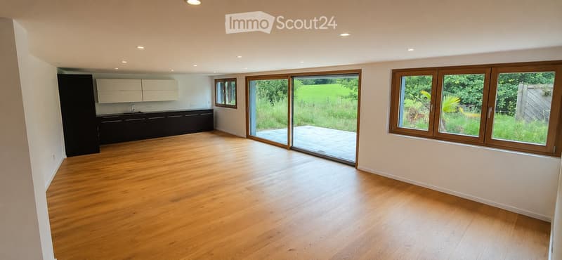 Bright, open-plan room with wooden flooring, white walls, and large windows and sliding doors leading to a garden. Kitchen area with cabinets and sink visible in the background.