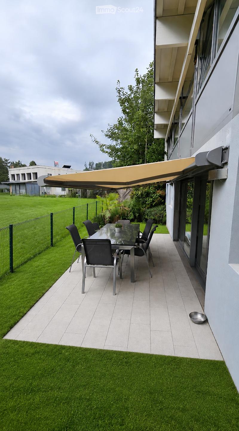 Outdoor patio with a glass dining table, six chairs, and a small potted plant, covered by a retractable awning attached to a modern building.