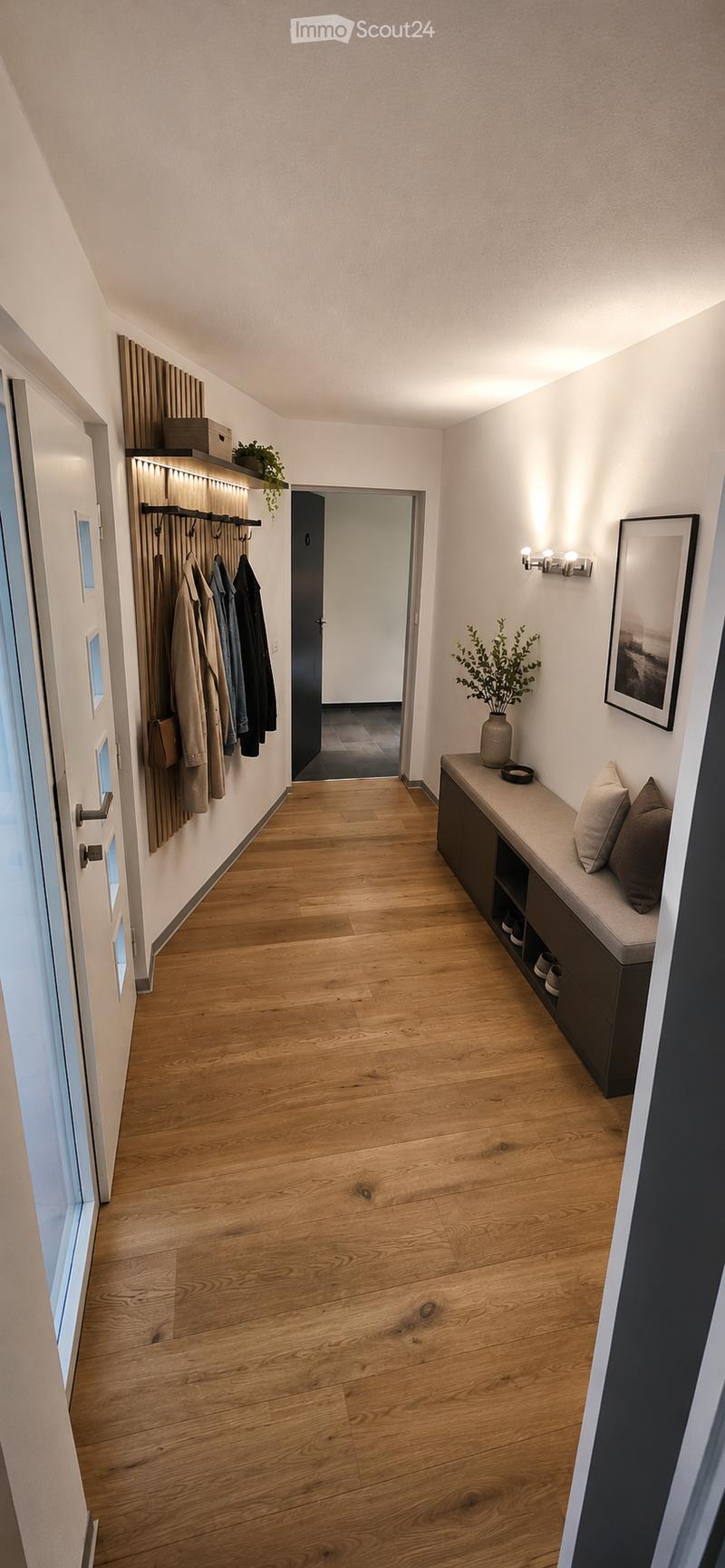 Modern hallway with wooden flooring, built-in bench with storage, coat rack, and decorative elements including a framed picture and potted plant.