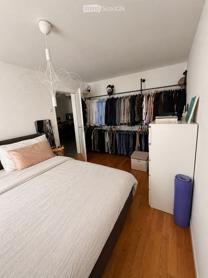 A well-lit bedroom with a large bed, pillows, a wardrobe with hanging clothes, a mirror, a lamp, and a yoga mat on the floor.