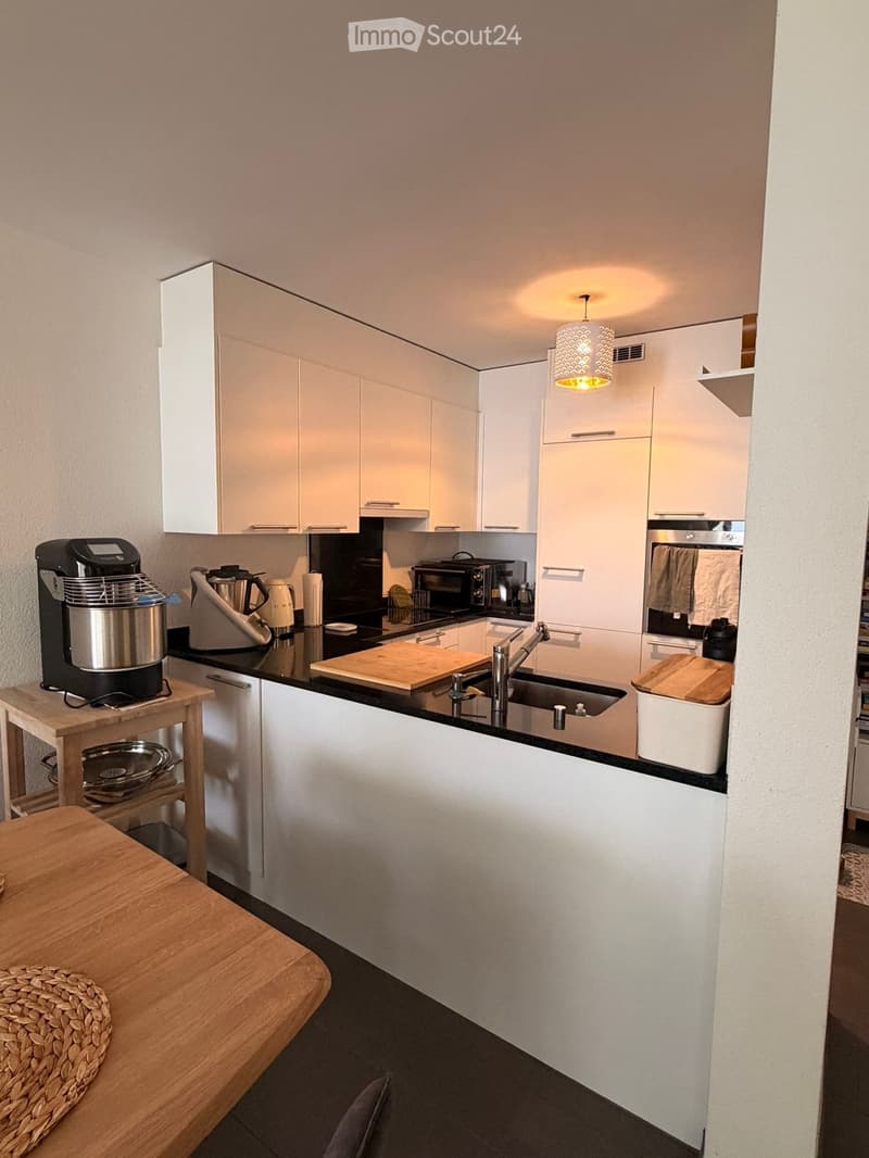 Modern kitchen with white cabinets, black countertops, and stainless steel appliances. Includes a mixer, microwave, cutting board, and a small wooden table with a woven basket.