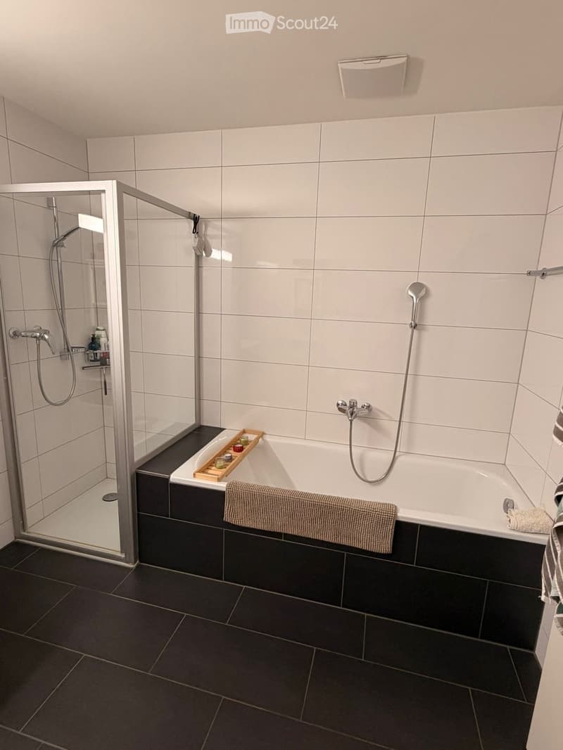 Modern bathroom with a white bathtub, glass shower enclosure, dark tiled floor, and a wooden tray with candles on the edge of the tub.