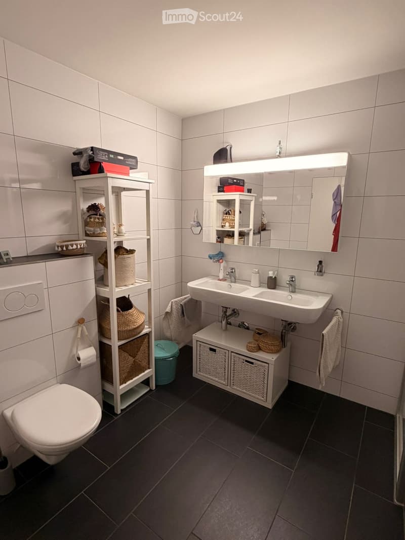 Modern bathroom with dark tiled floor, white tiled walls, double sink, large mirror with built-in lighting, white shelving unit with baskets, and a toilet.