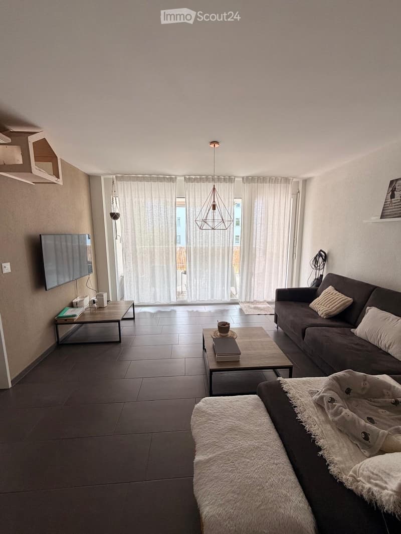 Modern living room with dark tiled floor, large window with white curtains, sofa with throw blanket, coffee table, and TV mounted on wall.